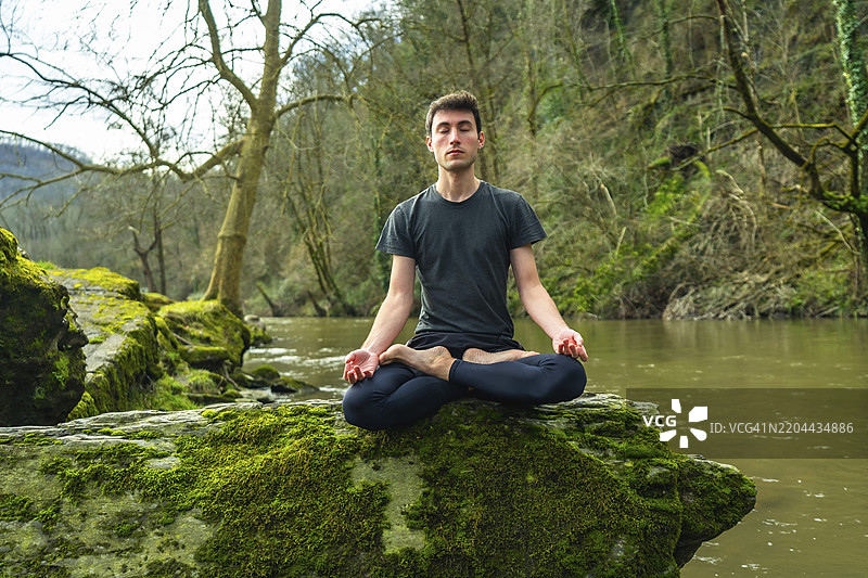 瑜伽教练坐在苔藓覆盖的岩石上，以莲花姿势冥想，周围是宁静的河流和郁郁葱葱的自然环境，传递正念与宁静。图片素材
