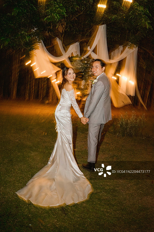 快乐的亚洲新婚夫妇在婚礼灯光下手牵手庆祝，笑着看向镜头图片素材