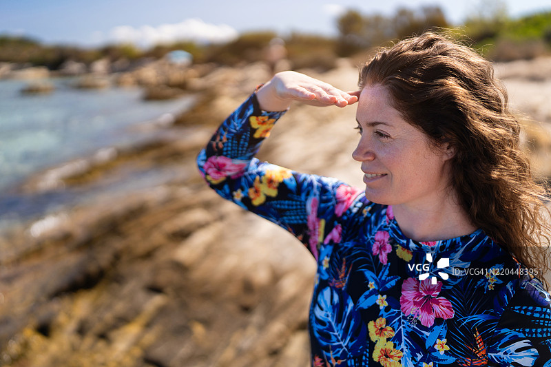 美丽的女人站在岩石海滩上，欣赏海景。她穿着潜水服，准备进行自由潜水。图片素材