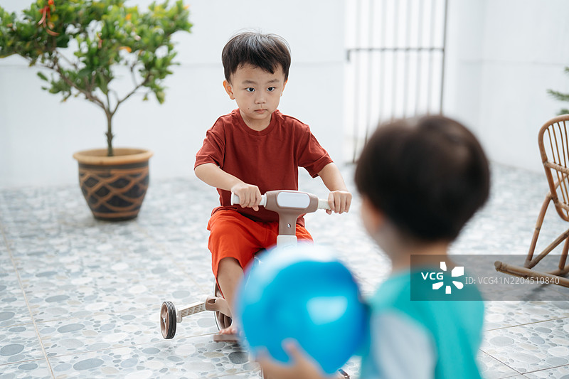 亚洲小男孩在奔跑，手里拿着球，而他的哥哥在骑自行车。图片素材