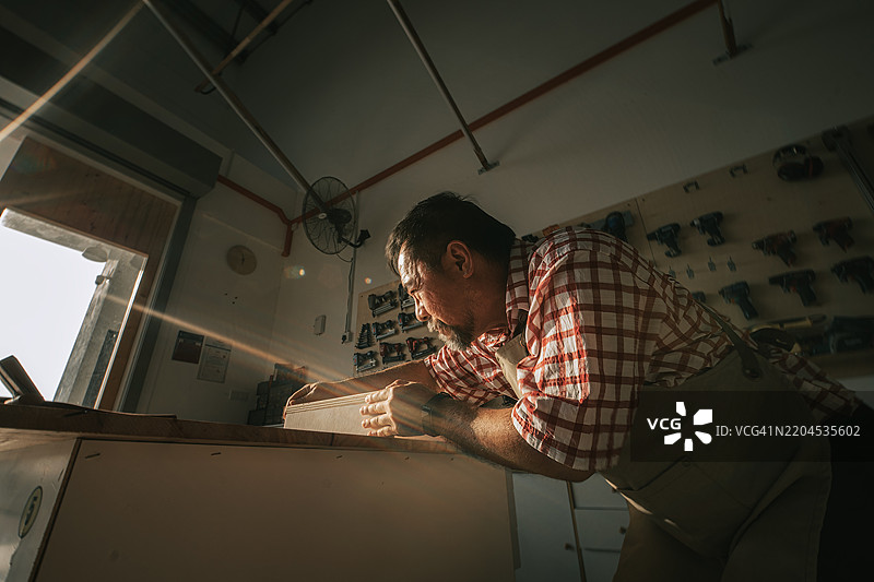 工艺精湛的亚洲华裔老木匠眯着眼睛测量木材，在木工车间工作图片素材