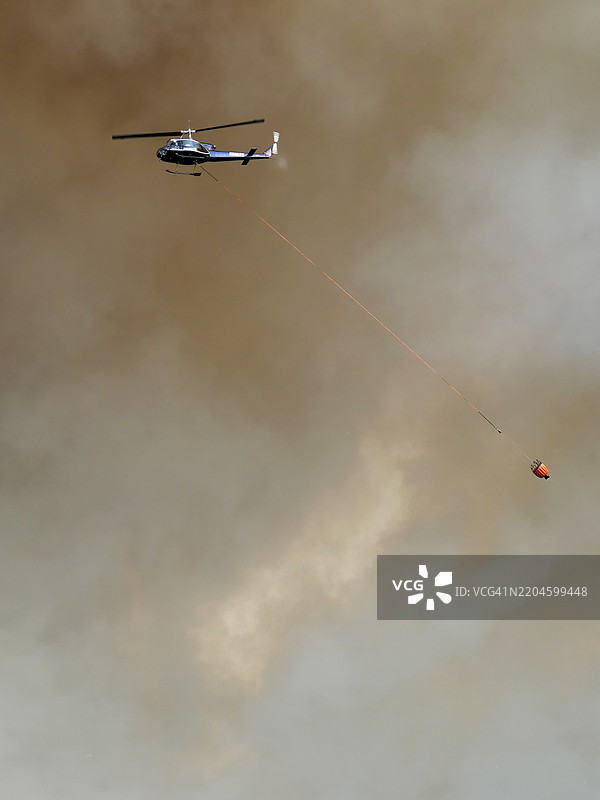 一架攻击直升机在森林火灾的烟雾前方飞行，吊着一个水桶。图片素材