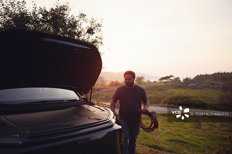 男子在晴朗的天空下走近汽车引擎盖，手中握着电缆。图片素材