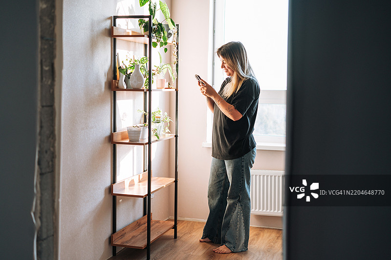 年轻女性在家中靠近摆满室内花卉的书架使用手机图片素材