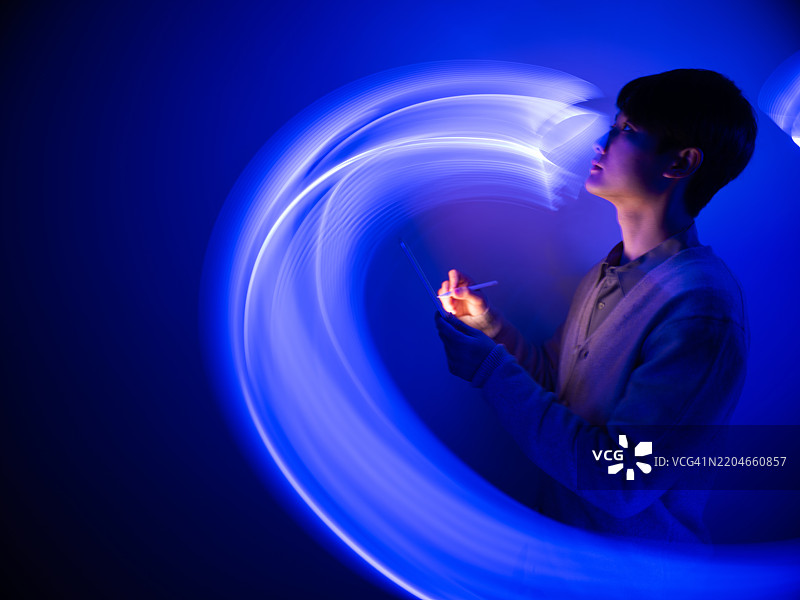 一位年轻男子在流动的光轨中使用数字平板电脑。仿佛置身于梦与现实之间的世界。人工智能概念，LED灯的长时间曝光。图片素材