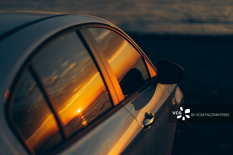 金色日落的倒影映在停靠在水边的银色汽车窗户上。温暖的阳光色调与车辆的冷金属色调形成对比。图片素材