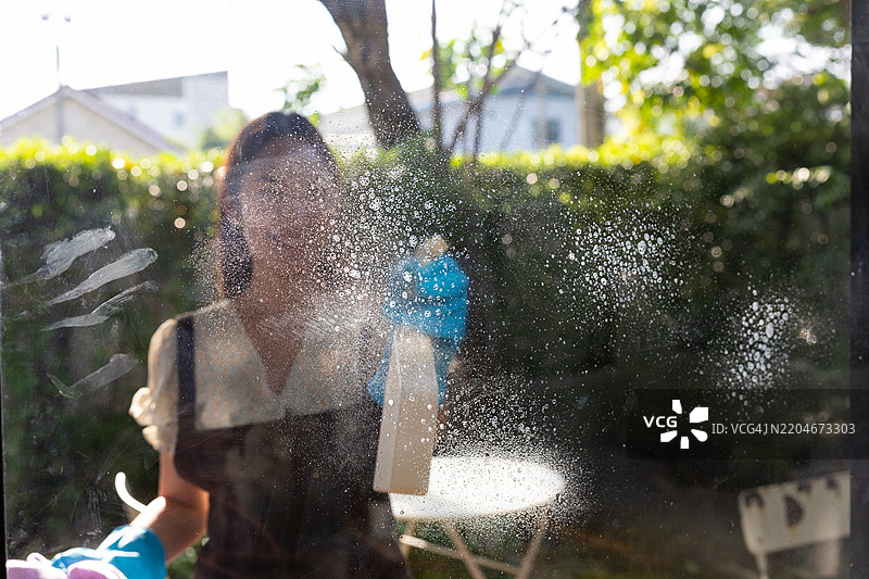 年轻的亚洲女性正在用清洁喷雾和布擦拭玻璃，确保家中无尘且闪亮，专注于日常家务维护，营造一个清新整洁的居住空间。图片素材
