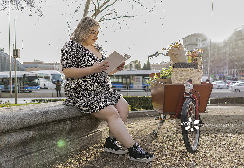 年轻女性在城市公园里阅读一本书，身旁是一辆装满植物和花卉的货运自行车。图片素材