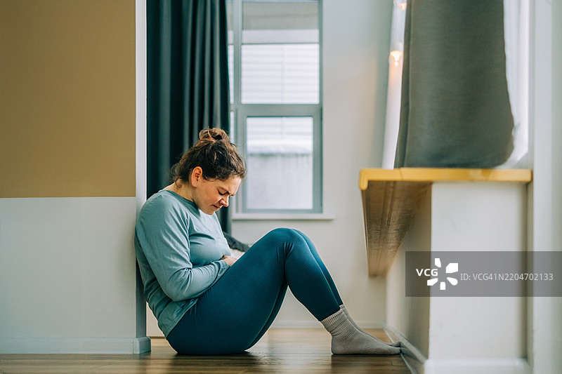 年轻女性因腹痛坐在家里的地板上图片素材