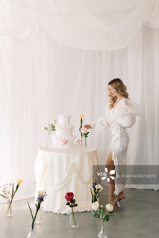 一位穿着优雅裙子的美丽女性站在一张摆满装饰蛋糕和甜点以及鲜花的桌子旁，周围是明亮而精致的春季室内环境。一位迷人的女性正在庆祝她的生日。新娘穿着婚纱，旁边是一块婚礼蛋糕。图片素材