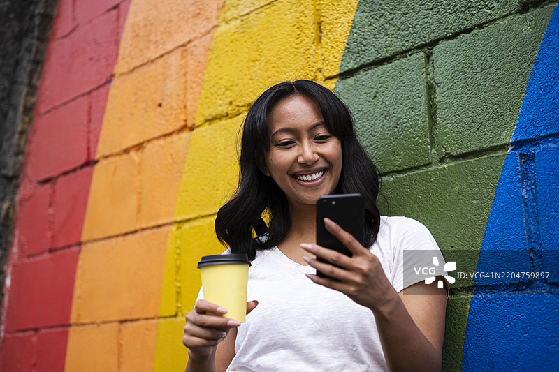 快乐的女人靠在多彩的墙上，手持咖啡杯使用智能手机图片素材