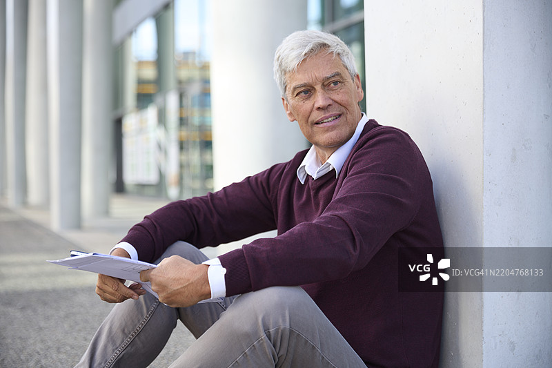 沉思的老年男子坐在建筑物旁边，手里拿着文件图片素材