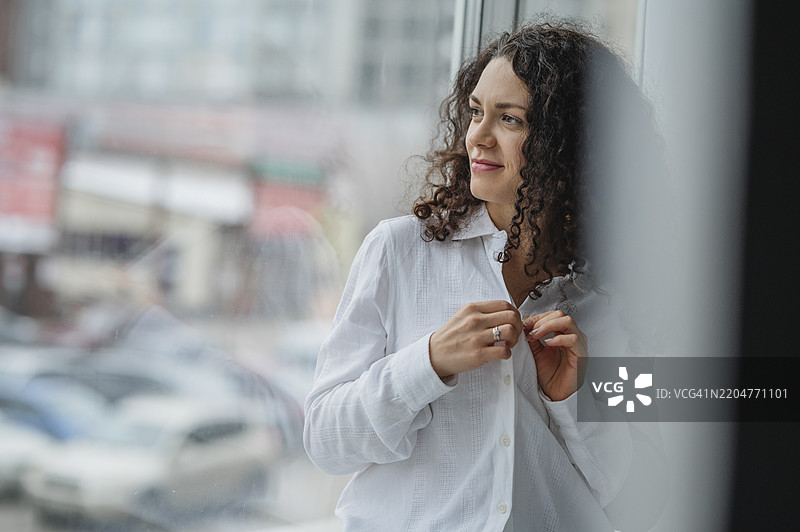 微笑的卷发女性在窗边扣衬衫钮扣图片素材