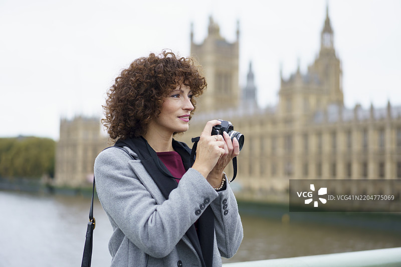 微笑的卷发女性在英国伦敦市议会大楼附近用相机拍照图片素材