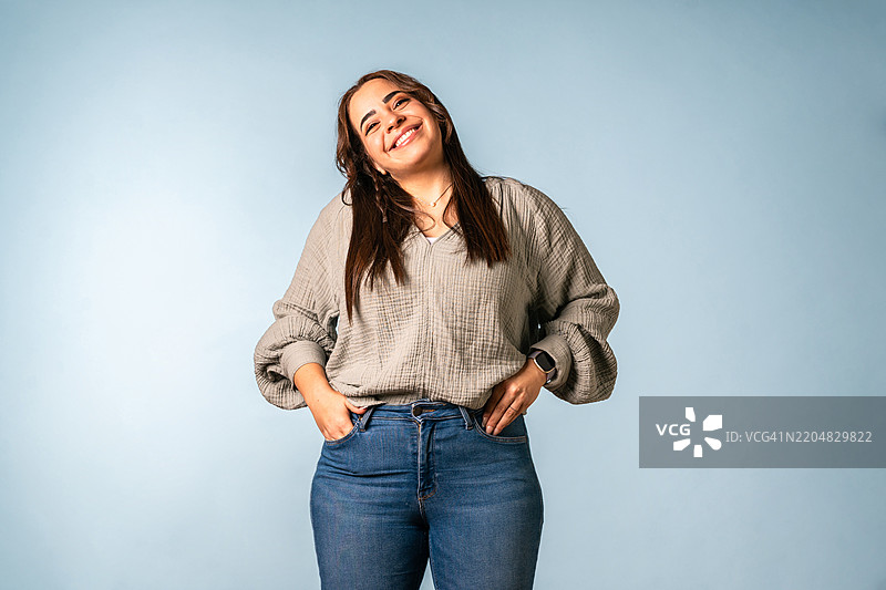 年轻的中东女性穿着休闲的米色毛衣和牛仔裤，在蓝色摄影棚背景前表达快乐。图片素材