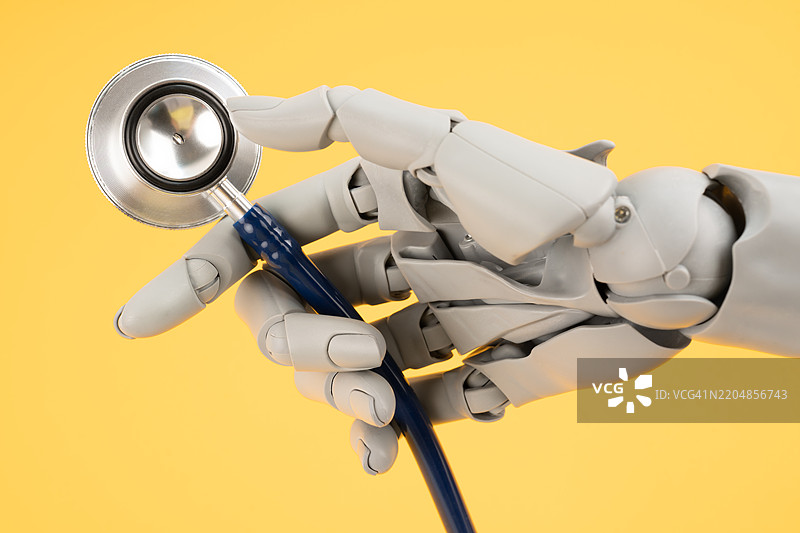 机器人手玩具手指握着医疗听诊器，背景为黄色，体现了人工智能和智能技术在医学和诊断中的概念。图片素材