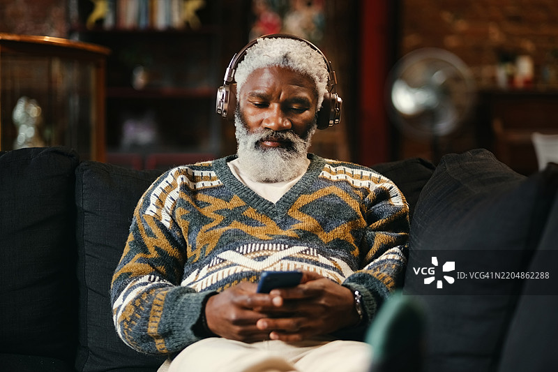在家中放松的男人，戴着耳机，手持智能手机，正在客厅享受播放列表。成熟的人在沙发上休息，享受音乐和音频科技的舒适体验。图片素材