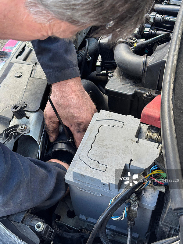 全景图像显示一位穿着蓝色工装的汽车修理工正在修理汽车发动机，手上沾满油污，打开的汽车引擎盖展示着发动机，修理工正在用塑料扎带拧紧进气管，画面聚焦于前景。图片素材