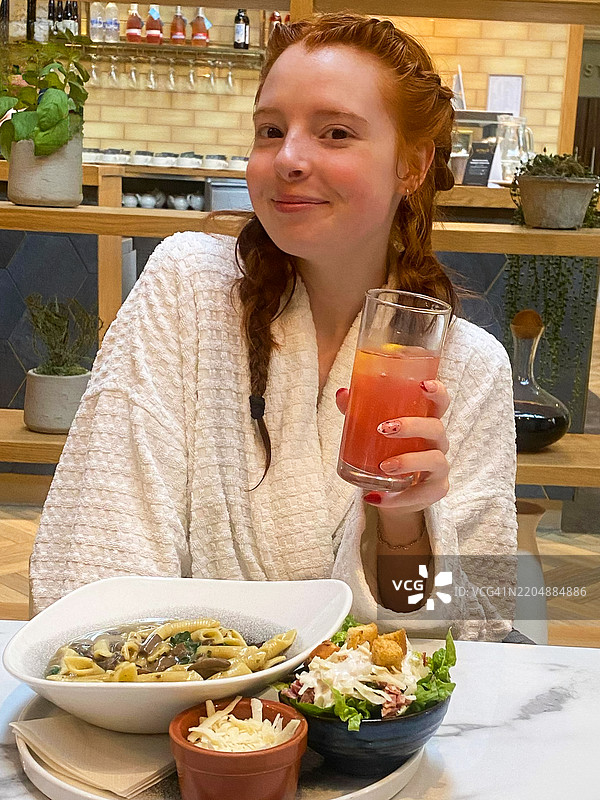 特写镜头：一位迷人的红发女性，扎着辫子，穿着白色水疗浴袍，手中拿着一杯软饮，旁边是一份蘑菇和婴儿菠菜意大利面的餐点，配有凯撒沙拉和碎奶酪，环境为桑拿和水疗餐厅。图片素材