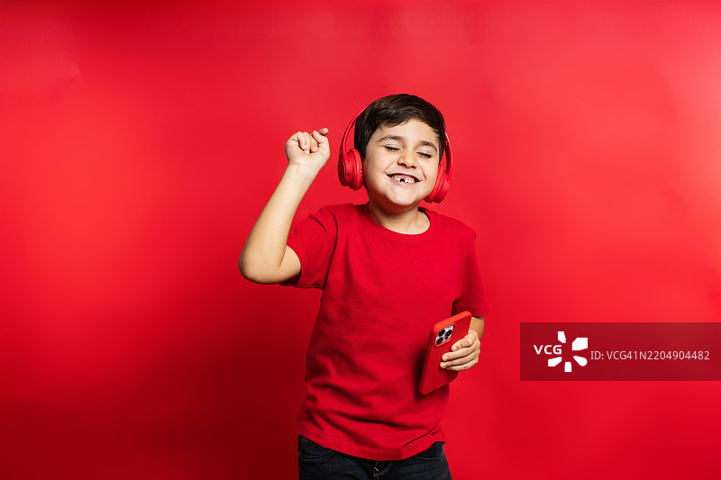 兴奋的孩子在红色背景下戴着耳机，享受着音乐，手里拿着智能手机。图片素材