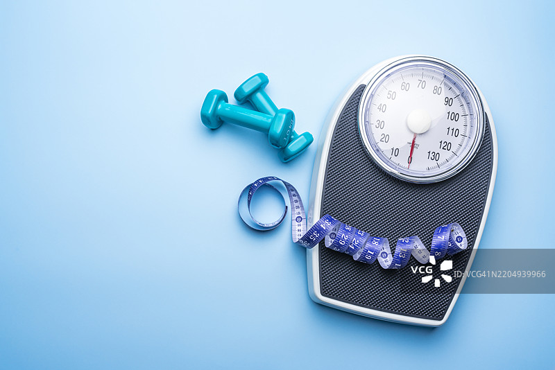 蓝色背景上的浴室秤、卷尺和哑铃，留有空白空间。体重控制、锻炼和健康概念。图片素材