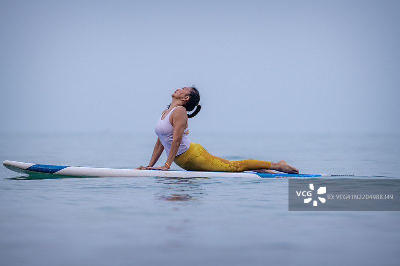 穿着白色背心和黄色紧身裤的女性在平静的海洋水面上练习瑜伽，优雅而专注地进行平衡动作，展现出核心力量的概念。图片素材