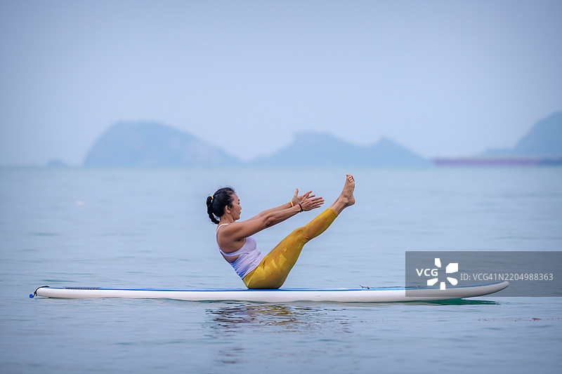 穿着白色背心和黄色紧身裤的女性在平静的海水中练习瑜伽，站在冲浪板上。她优雅而专注地进行平衡练习，展现出核心力量的概念。图片素材