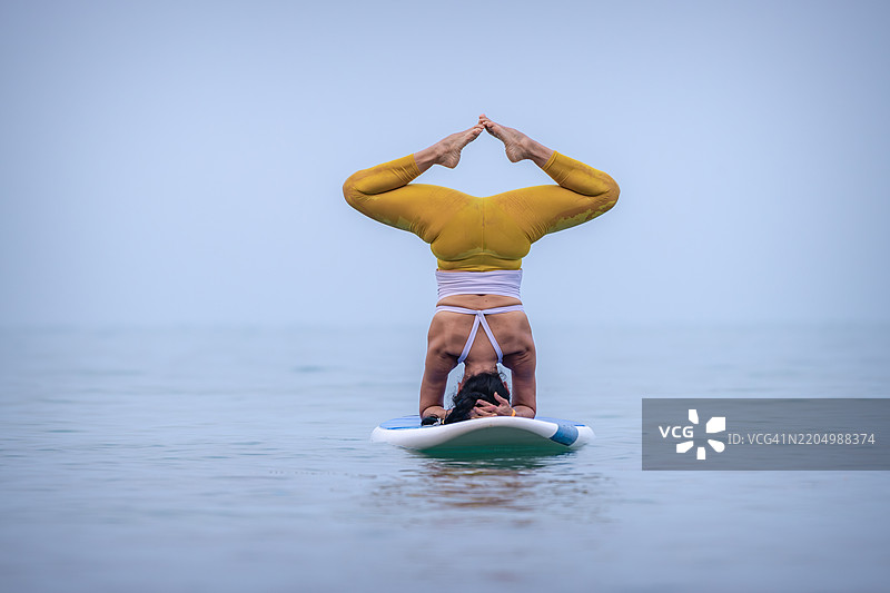 穿着白色背心和黄色紧身裤的女性在平静的海面上练习瑜伽，优雅地进行平衡动作，展现出专注和核心力量的概念。图片素材