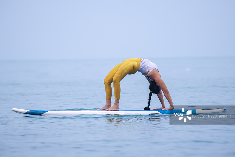 穿着白色背心和黄色紧身裤的女性在平静的海洋水面上练习瑜伽，优雅而专注地进行平衡动作，展现核心力量的概念。图片素材