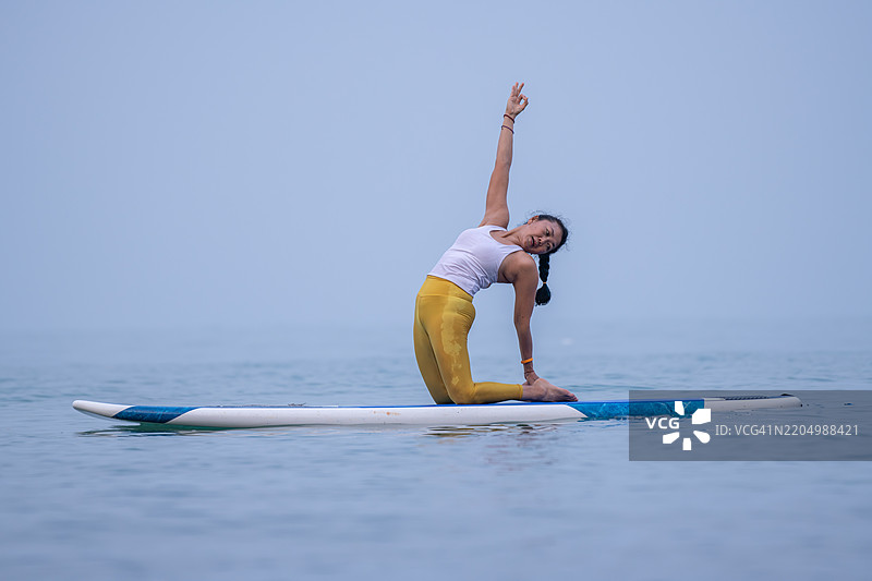 穿着白色背心和黄色紧身裤的女性在平静的海洋水面上练习瑜伽，优雅地进行平衡动作，展现出专注和核心力量的概念。图片素材