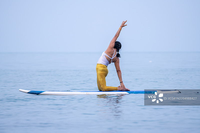 穿着白色背心和黄色紧身裤的女性在平静的海洋水面上练习瑜伽，优雅地进行平衡动作，展现出专注和核心力量的概念。图片素材
