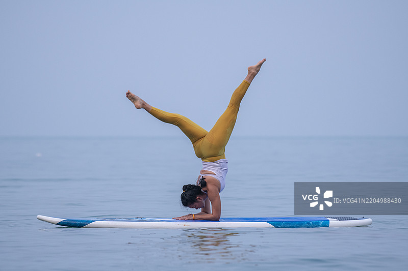 穿着白色背心和黄色紧身裤的女性在平静的海面上练习瑜伽，优雅地进行平衡动作，展现出专注和核心力量的概念。图片素材