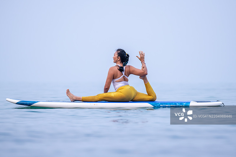 穿着白色背心和黄色紧身裤的女性在平静的海洋水面上练习瑜伽，优雅地进行平衡动作，展现出专注和核心力量的概念。图片素材
