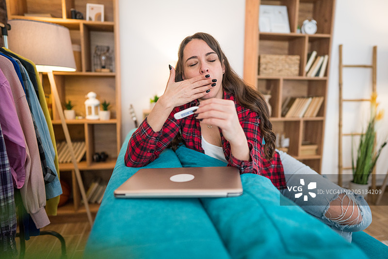 一位疲惫的女性打着哈欠，手中拿着温度计。图片素材