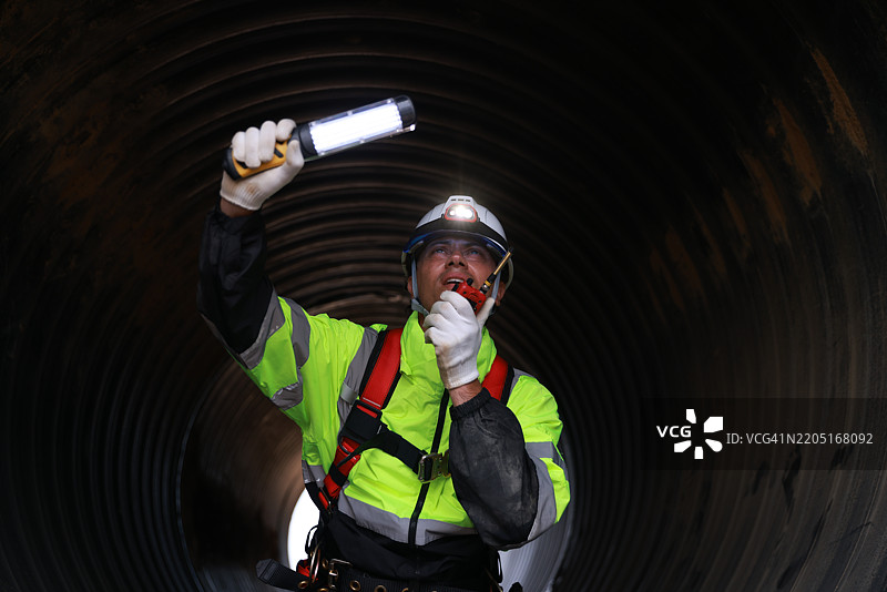 一名穿着安全带和安全帽的建筑工人正在工地上检查一根大型排水管，身边有一个工具箱。图片素材