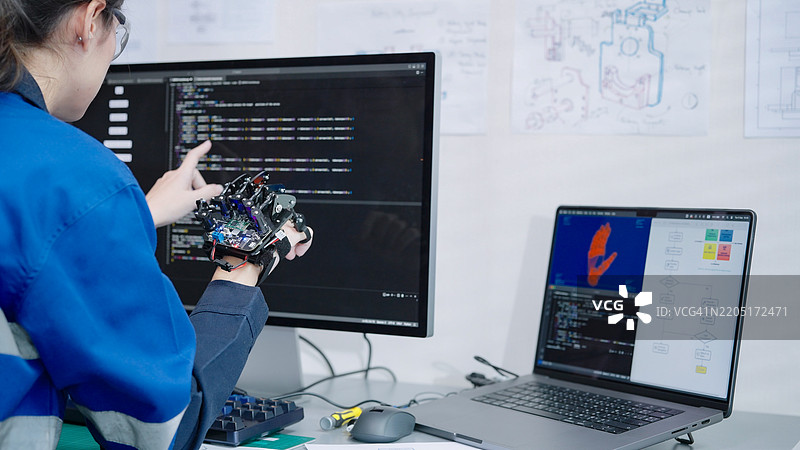 女性工程师开发者在计算机上编写代码，使用机器人手臂模型原型控制电机控制传感器进行测试，涉及机械工程和软件开发。图片素材