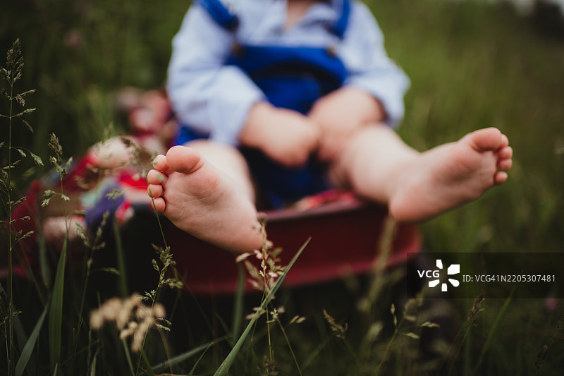 小男孩赤脚坐在红色小车里，脚上沾满了泥土。图片素材