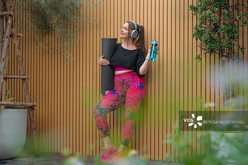 穿着运动服的美丽大码女性，手持瑜伽垫和可重复使用的水瓶，戴着耳机。图片素材