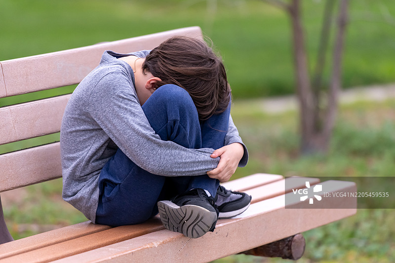 沮丧的哭泣男孩坐在公园长椅上捂住脸。图片素材