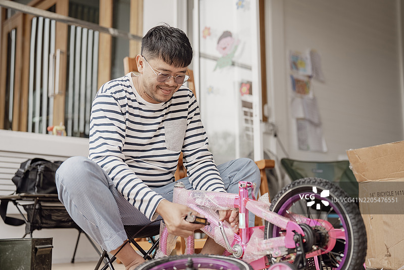 积极情绪的亚洲父亲穿着条纹衬衫，坐在椅子上用工具组装一辆粉色自行车，旁边有包装材料。图片素材