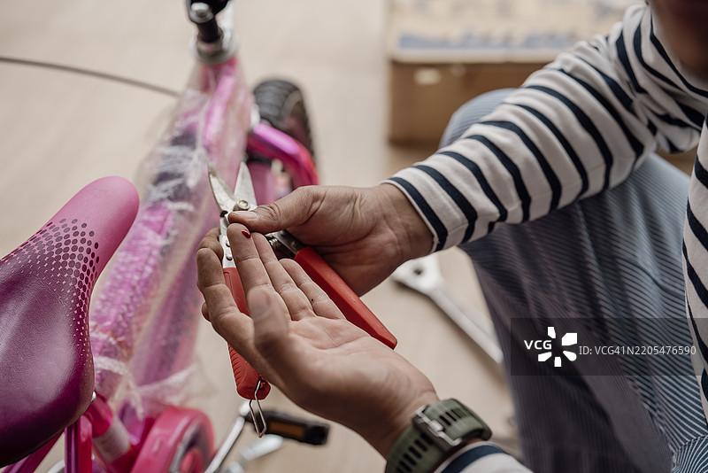 一名穿着条纹衬衫的男子在组装一辆粉色自行车时，检查手上的小伤口，同时手里拿着红色手柄的钳子。图片素材