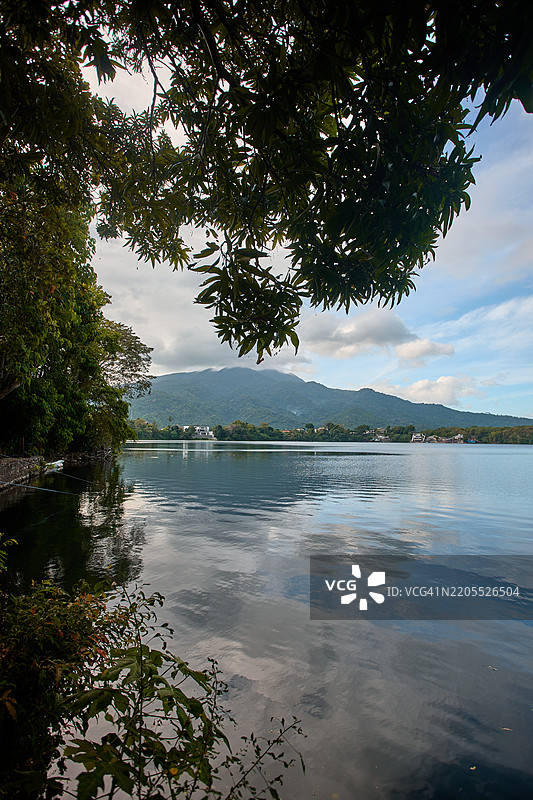 鳄鱼湖是一个小型火山口湖，前景被郁郁葱葱的树叶环绕，背景是位于拉古纳洛斯巴尼奥斯的马基林山的起伏山丘。图片素材