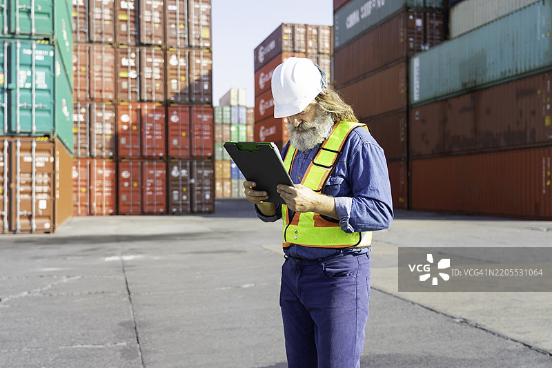 仓库主管专注于用夹板监控库存，戴着安全帽和反光背心。图片素材