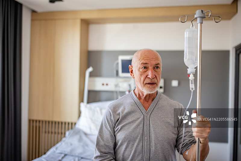 一位老年男性在医院病房接受医疗护理，手握静脉输液架，神情沉思。图片素材