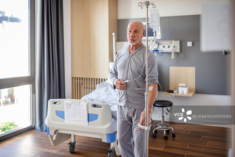 老年男子站在医院病床旁，接受医疗专业人员的护理和支持，正在康复中。图片素材