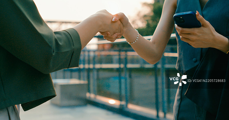 两位年轻的亚洲女商人在城市环境中握手，象征着成功的交易或合作，展现可持续的户外商业。图片素材