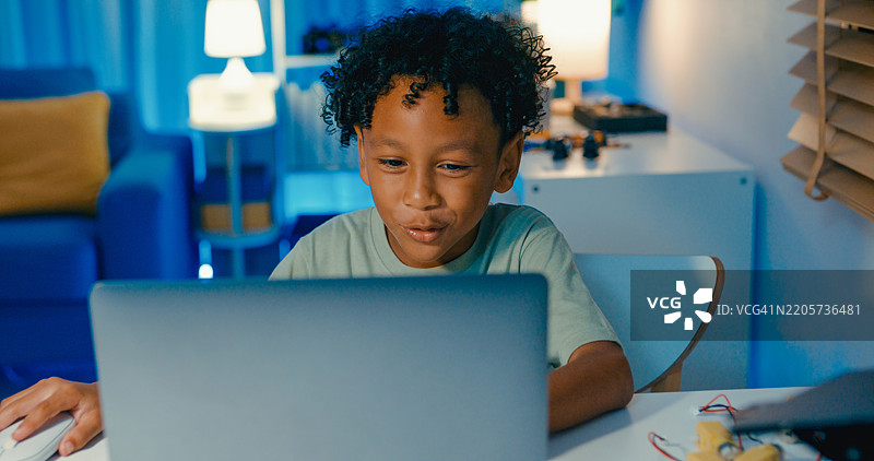 年轻的非裔美国男孩在家中夜晚使用笔记本电脑学习编程代码，参与与机器人相关的STEM学习项目。在线STEM教育机器人课程。图片素材