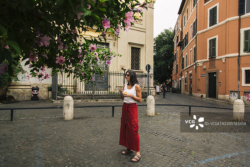 色彩缤纷的街道上，一位女性手持水果沙拉，周围环绕着盛开的花朵。图片素材