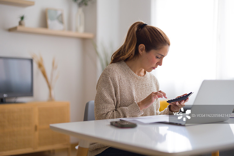 年轻女性在家中使用计算器和笔记本电脑计算家庭账单图片素材