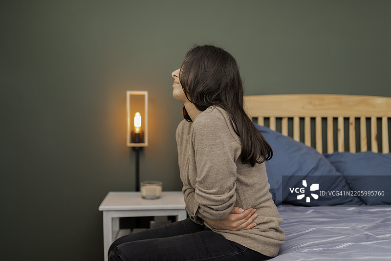 年轻女性坐在床上，感受腹部疼痛，周围是温馨的卧室环境图片素材